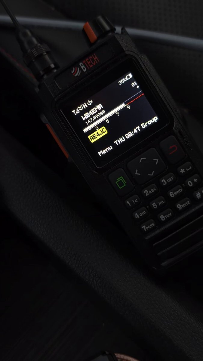 A BTECH handheld radio in a car center console at night, screen lit showing contact WB4EMR on 147.090 MHz and callsign AE4JC in the group list