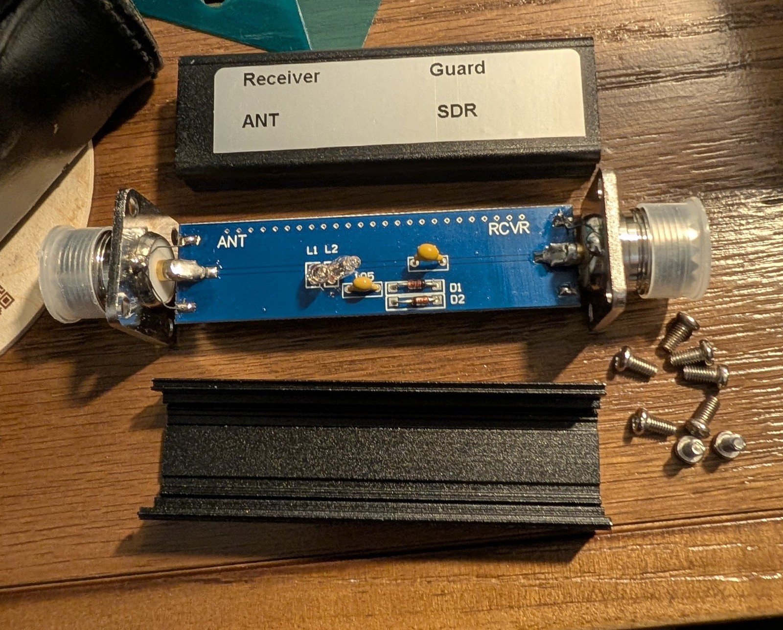 A disassembled SDR receiver guard circuit board labeled ANT and RCVR, next to its aluminum enclosure and mounting screws