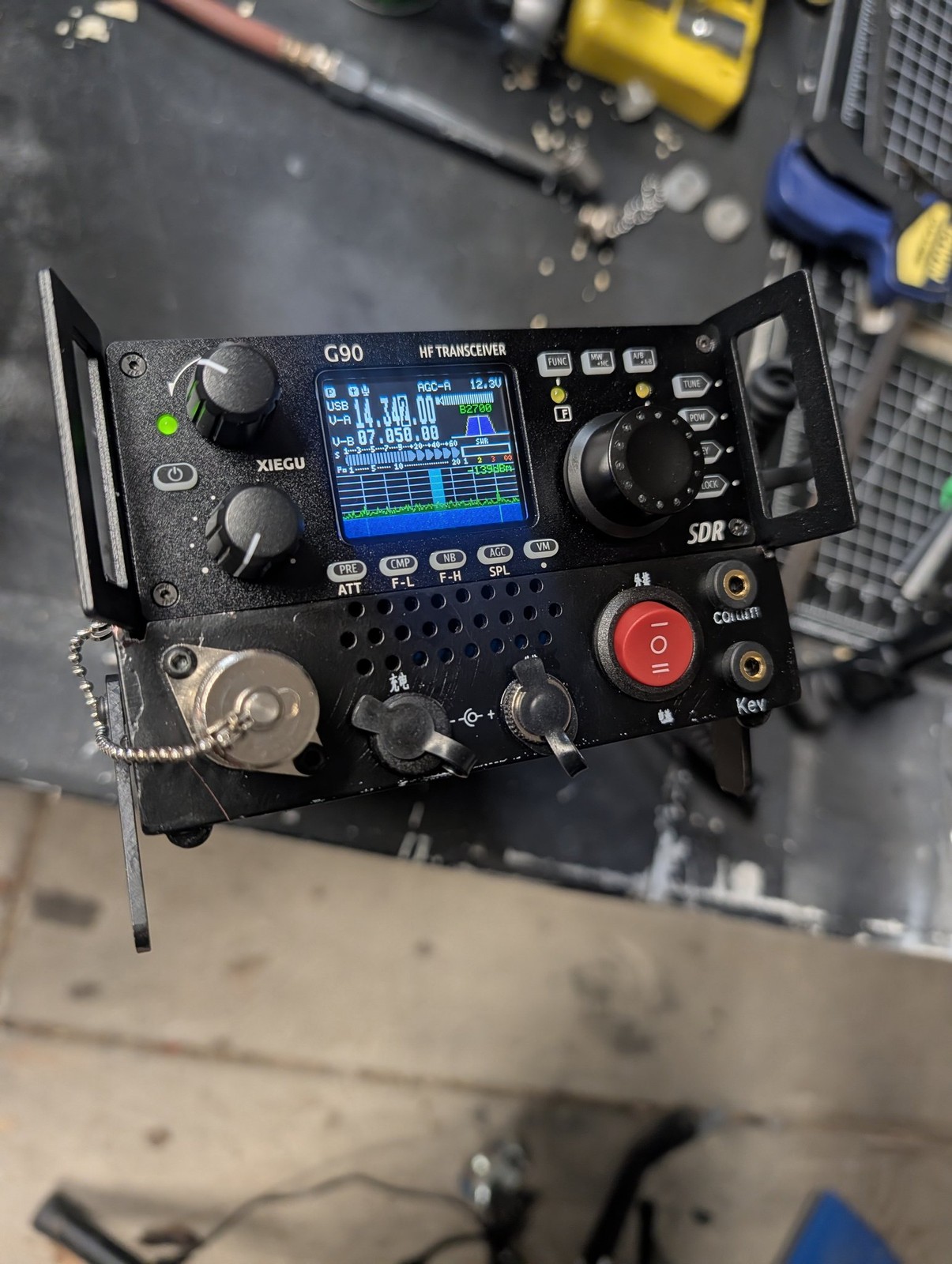 A Xiegu G90 HF transceiver with a newly added external battery pack and stand, powered on and showing 14.340.00 MHz USB on its display, sitting on a workbench